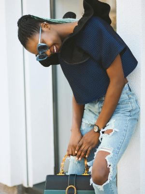 Fashion-forward woman smiling in blue knit top and sunglasses, holding a bag.